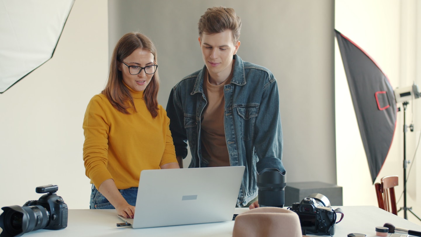 Zwei Fotografen besprechen einen Laptop am Tisch in einem Studio.